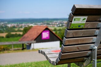 Holzbank mit Rückenlehne im Vordergrund, darauf ein Aufkleber "KinderErlebnisWeg", im Hintergrund ein weiter Ausblick über die Hügellandschaft und ein kleines Häuschen mit lila Infotafel – aufgenommen bei sommerlichem Wetter in Oberderdingen. | © Kraichgau-Stromberg Tourismus e.V.