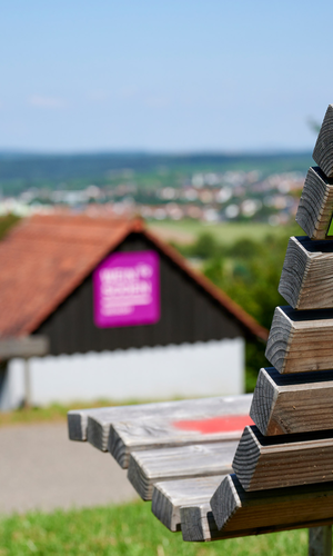 Holzbank mit Rückenlehne im Vordergrund, darauf ein Aufkleber "KinderErlebnisWeg", im Hintergrund ein weiter Ausblick über die Hügellandschaft und ein kleines Häuschen mit lila Infotafel – aufgenommen bei sommerlichem Wetter in Oberderdingen. | © Kraichgau-Stromberg Tourismus e.V.