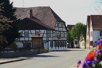 Fachwerkhaus mit rotem Ziegeldach und Schild 'Zum Löwen' an einer ruhigen Straße, rechts weitere Häuser und Bäume. | © Landratsamt Rhein-Neckar-Kreis