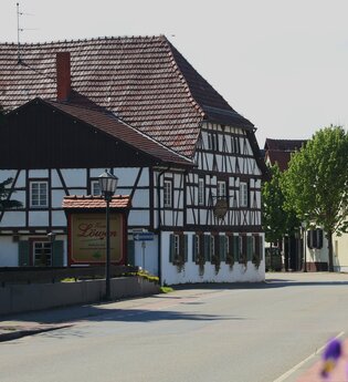 Fachwerkhaus mit rotem Ziegeldach und Schild 'Zum Löwen' an einer ruhigen Straße, rechts weitere Häuser und Bäume. | © Landratsamt Rhein-Neckar-Kreis