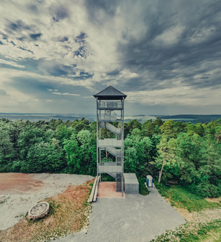 Luftaufnahme vom Aussichtsturm mit Nebengebäude auf dem Stahlberg in Külsheim-Uissigheim. Im Hintergrund geht der Blick ins bewaldete Taubertal.  Der Himmel zeigt ein wildes Wolkenschauspiel in dunkelblau und weiß. | © Liebliches Taubertal