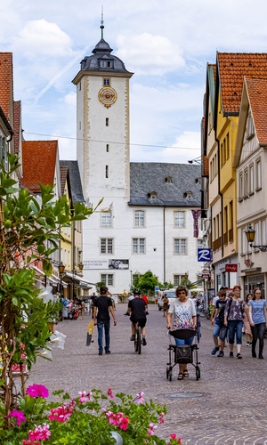 Blick in die Fußgängerzone in der Innenstadt von Bad Mergentheim. Es  sind viele Menschen unterwegs. Im Hintergrund ist das Residenzschloss und im Vordergrund Blumenkübel mit Sommerbepflanzung zu sehen.