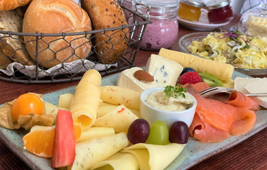 Teller mit Käse und Lachs. Im Hintergrund sind Brötchen und Marmelade zu sehen.