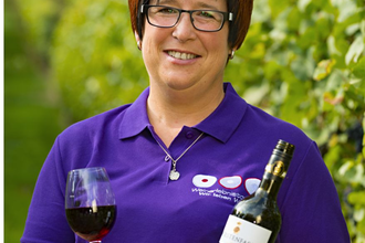 Eine Frau hält eine Flasche Rotwein und ein Glas in der Hand, sie steht in einem Weinberg | © Touristikgemeinschaft Hohenlohe e. V. | Obst- und Weinbau Dietz