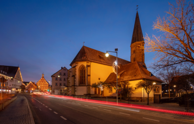 Abendaufnahme einer beleuchteten Kirche an einer Straße mit Lichtspuren von Autos. | © Stadt Gaildorf