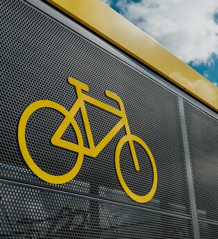 Auf dem Bild ist ein Ausschnitt der Fahrradabstellanlage von außen zu sehen, mit einem gelb aufgedruckten Fahrrad-Symbol.