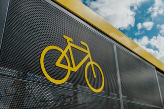 Auf dem Bild ist ein Ausschnitt der Fahrradabstellanlage von außen zu sehen, mit einem gelb aufgedruckten Fahrrad-Symbol.