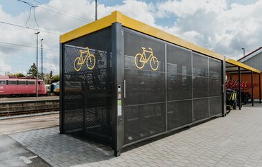 Auf dem Bild ist die gesamte Fahrradabstellanlage von außen zu sehen. Im Hintergrund ein Teil des Bahnhofs.