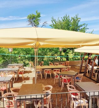 Sonnige Außenterrasse eines Cafés mit Tischen, Stühlen und großen Sonnenschirmen, umgeben von grünen Bäumen