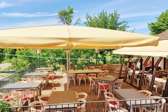 Sonnige Außenterrasse eines Cafés mit Tischen, Stühlen und großen Sonnenschirmen, umgeben von grünen Bäumen