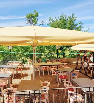 Sonnige Außenterrasse eines Cafés mit Tischen, Stühlen und großen Sonnenschirmen, umgeben von grünen Bäumen