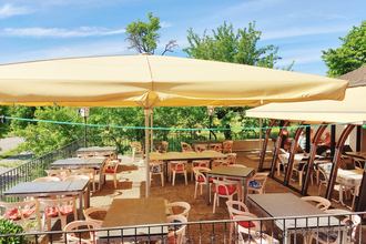 Sonnige Außenterrasse eines Cafés mit Tischen, Stühlen und großen Sonnenschirmen, umgeben von grünen Bäumen