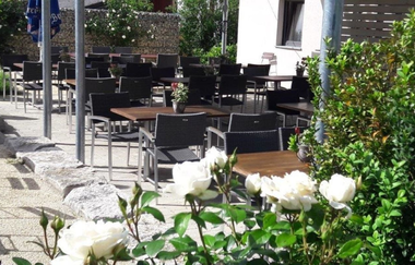 hell gepflasterte Terrasse mit braunen Tischen und schwarzen Stühlen mit Armlehne im Vordergrund rechts ein grüner Busch und weiß blühende Rosen | © Gasthof Kocherbähnle GmbH