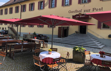 Der Biergarten vor dem Gasthof ist rustikal mit Tischen und Stühlen und roten Sonnenschirmen bestückt | © Touristikgemeinschaft Hohenlohe e. V. | Gasthof zur Post