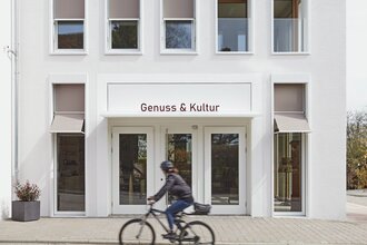 Außenansicht des weißen Gebäudes von Genuss & Kultur in Güglingen, FahrradfahrerIn davor | © NZT
