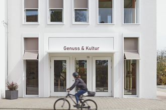 Außenansicht des weißen Gebäudes von Genuss & Kultur in Güglingen, FahrradfahrerIn davor | © NZT