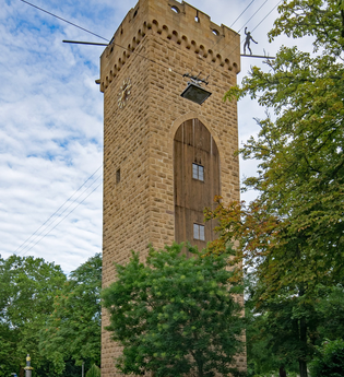 Im Fokus steht ein hoher Turm aus Naturstein. Auf einer Seite sind die Steine durch eine bogenförmigen Holzkonstruktion mit Fenster unterbrochen. Auf einer weiteren Seite ist eine Uhr zu sehen. Vor dem Turm sind auf dem Gehweg Bäume gepflanzt