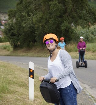 Lächelnde Frau fährt auf einem Segway auf der Straße. Im Hintergrund zwei weitere Personen auf einem Segway. | © GötzMotion