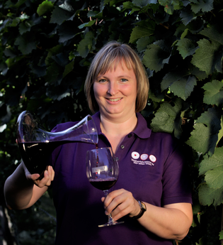 Weinerlebnisführerin Gudrun Ungerer steht vor Reben mit einem Glas Rotwein in der Hand und schenkt Rotwein aus einem Dekanter ein. | © Touristikgemeinschaft Hohenlohe e.V. | Weingut Ungerer