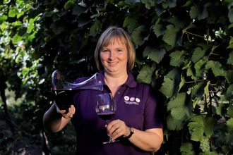 Weinerlebnisführerin Gudrun Ungerer steht vor Reben mit einem Glas Rotwein in der Hand und schenkt Rotwein aus einem Dekanter ein. | © Touristikgemeinschaft Hohenlohe e.V. | Weingut Ungerer