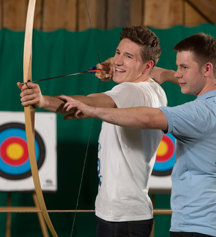 Ein Mann wird beim Indoor-Bogenschiessen von einem Heffner Outdoor Events Mitarbeiter eingewiesen | © Touristikgemeinschaft Hohenlohe e.V. | Heffner Outdoor Events
