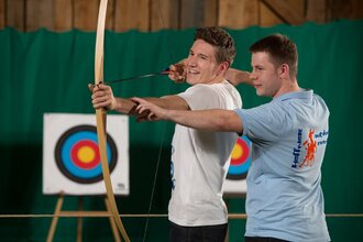 Ein Mann wird beim Indoor-Bogenschiessen von einem Heffner Outdoor Events Mitarbeiter eingewiesen | © Touristikgemeinschaft Hohenlohe e.V. | Heffner Outdoor Events