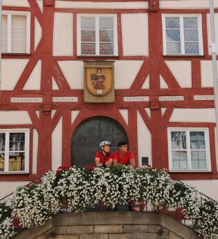 Zwei Personen stehen auf dem Treppenaufgang zum historischen Rathaus mit rotem Fachwerk | © Touristikgemeinschaft Hohenlohe e. V. | Andi Schmid