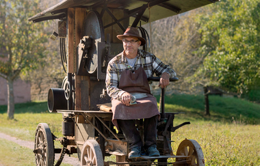 Ein Mann sitzt auf einer alten Landmaschine im Freien, umgeben von Bäumen und Wiesen. | © Hohenloher Freilandmuseum