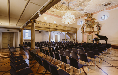 Ein eleganter Festsaal mit Stuhlreihen, einem Flügel, verzierten Wänden und einer bemalten Decke. | © NICO KURTH