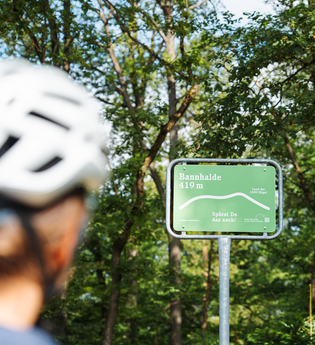 Eine Radfahrerin mit Helm blickt auf ein grünes Schild an der Bannhalde (419 m) im Naturpark Stromberg-Heuchelberg mit der Aufschrift „Spürst Du das auch?“. | © SaddleStories.at
