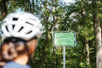 Eine Radfahrerin mit Helm blickt auf ein grünes Schild an der Bannhalde (419 m) im Naturpark Stromberg-Heuchelberg mit der Aufschrift „Spürst Du das auch?“. | © SaddleStories.at