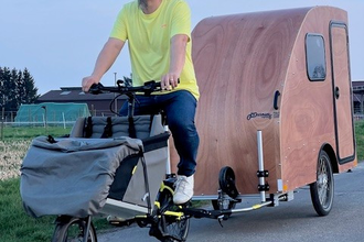 Ein Mann mit Jeans und gelbem T-Shirt bekleidet fährt auf einer asphaltierten Strasse mit einem Lastenfahrrad an dem ein kleiner Fahrradwohnwagen aus Holz angehängt ist | © Dennis Hirschmann
