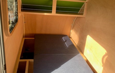 Fahrradwohnanhänger von innen. Die Innenwände sind aus Holz. Auf der linken Seite ist ein kleines Fenster. Der gebogene Dachhimmel ist grün gestrichen. Die Sitzbank kann zu einem Bett umgebaut werden und  bietet eine Liegefläche von 90 x 2 m | © Dennis Hirschmann