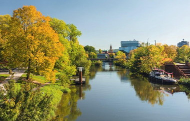 Das Bild zeigt den ruhigen Neckar, umgeben von Bäumen im Herbst. Die spiegelnde Wasseroberfläche reflektiert die Bäume und Gebäude im Hintergrund. Am Flussufer sind einige Boote zu sehen, die friedliche Szene wird von strahlendem Sonnenschein beleuchtet.