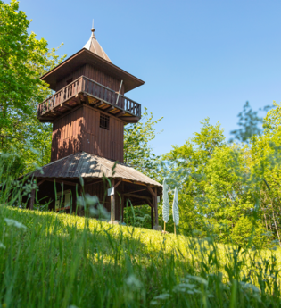 Ein hölzerner Aussichtsturm steht auf einer Wiese, umgeben von Bäumen und blühenden Blumen, unter einem klaren blauen Himmel. | © Stadt Gaildorf