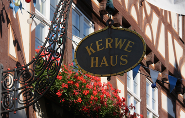 Das Schild des Kerwehauses, ein schwarzes Oval mit goldener Schrift, an der Fachwerkfassade, im Hintergrund blühen rote Geranien.