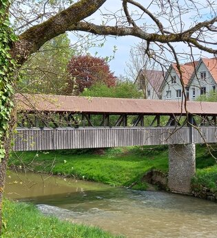 Holzbrücke über einen Fluss in Gaildorf, umgeben von Bäumen und Fachwerkhäusern. | © Petra Natzkowski