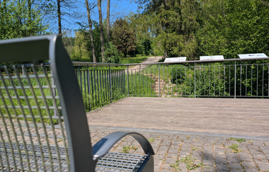Im Vordergrund eine Metallbank auf einem Holzdeck. Im Hintergrund sind Infotafeln zu der historischen Stätte "Leberbrunnen" zu erkennen. | © Touristikgemeinschaft HeilbronnerLand