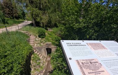 Nahaufnahme einer Infotafel am Leberbrunnen. Im Hintergrund ist die in Sandstein eingefasste Quelle zu sehen. | © Touristikgemeinschaft HeilbronnerLand