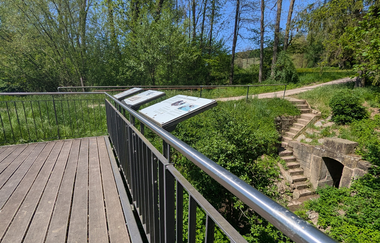 Bild vom Holzdeck mit Infotafeln am Leberbrunnen. Im Hintergrund ist die historische Brunnenanlage mit Sandsteineinfassung zu erkennen. | © Touristikgemeinschaft HeilbronnerLand