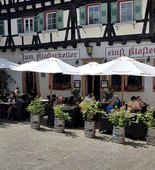 Gastronomische Außenbestuhlung vor Fachwerkhaus mit weißen Sonnenschirmen, Gästen und Fass-Pflanzkübeln, Schriftzug „Zum Klosterkeller“ - einst Klosterschmiede" an der Fassade. | © Park-Gastronomie