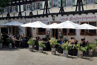 Gastronomische Außenbestuhlung vor Fachwerkhaus mit weißen Sonnenschirmen, Gästen und Fass-Pflanzkübeln, Schriftzug „Zum Klosterkeller“ - einst Klosterschmiede" an der Fassade. | © Park-Gastronomie