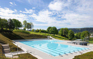 Ein großes Freibad mit klarem Wasser, umgeben von grünen Hügeln und Bäumen unter einem blauen Himmel. | © Stadt Gaildorf