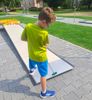 Ein Junge steht mit seinem Golfschläger vor der Minigolf-Bahn; im Hintergrund ist ein Park mit Wiese und Bäumen zu sehen. | © Touristikgemeinschaft Odenwald e.V.