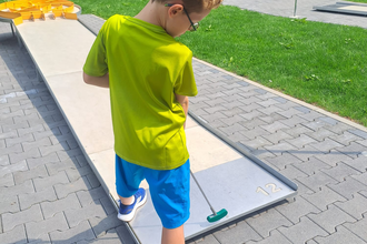 Ein Junge steht mit seinem Golfschläger vor der Minigolf-Bahn; im Hintergrund ist ein Park mit Wiese und Bäumen zu sehen. | © Touristikgemeinschaft Odenwald e.V.