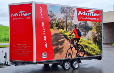 Stehender Fahrradanhänger mit einem Bild von einem Radfahrer am Fluss und das Logo von der Firma Müller Reisen. | © Müller Reisen Massenbachhausen
