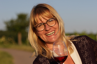 Portrait von Nicole Halter mit einem Glas Rotwein in der Hand. Mit einem gewinnenden Lächeln strahlt sie mit der Sonne um die Wette. | © DieWeinschmeckerin Nicole Halter