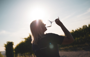 Ein Frau mit längeren, blonden Haaren die ein Glas Weißwein trinkt im Gegenlicht aufgenommen. Schöne Lichtstimmung im Sonnenuntergang mit ein Weinbergen im Hintergrund. | © Nicole Halter