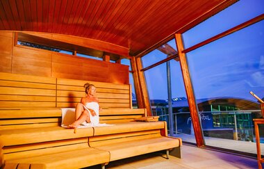Dame mit Handtuch sitzt in einer Sauna mit Blick auf das Gebäude des parc vitals | © Stadtwerke Crailsheim