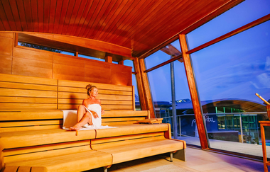 Dame mit Handtuch sitzt in einer Sauna mit Blick auf das Gebäude des parc vitals | © Stadtwerke Crailsheim